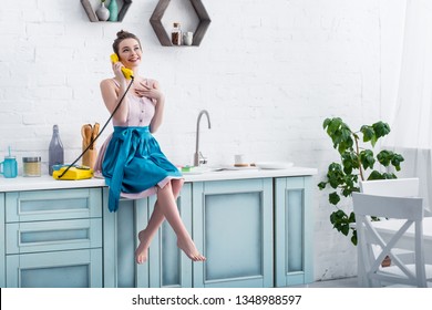 happy young woman in apron sitting on table and talking on retro yellow telephone in kitchen  