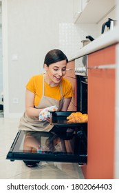 Happy Young Woman In Apron Squatting Down In The Kitchen Near The Open Oven And Admiring Delicious Croissants On A Baking Pan