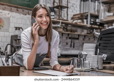 Happy young woman bar owner talking on smartphone at the counter and looking away smiling. Close up shot of attractive cheerful barista, cafe worker, waitress taking noting checking online order - Powered by Shutterstock