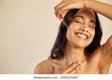 Happy young woman with bare shoulder applying serum on face with closed eyes. Beautiful hispanic young woman moisturizes her skin with serum isolated against beige background with copy space.
