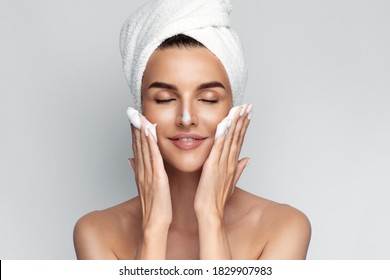 Happy young woman with bath towel on her head takes care of her skin face, applies cleansing foam after shower, smiling and closed eyes, isolated on grey background. Face wash. 