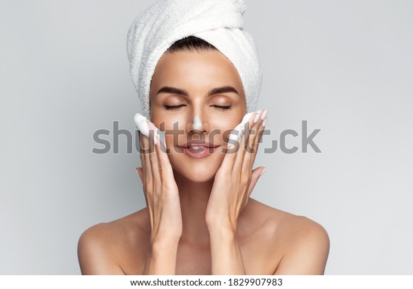 Happy young\
woman with bath towel on her head takes care of her skin face,\
applies cleansing foam after shower, smiling and closed eyes,\
isolated on grey background. Face wash.\

