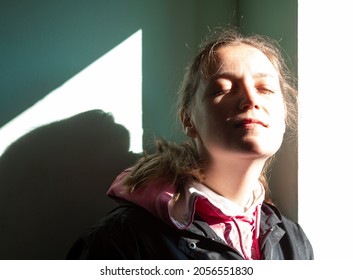 Happy young woman in black raincoat against wall basking in sun autumn day. Smiling girl with closed eyes standing near door sunrays. Lifestyle real people Seasonal depression mental health, Vitamin D
