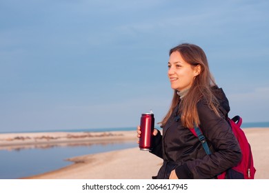 Happy Young Woman In Black Raincoat With Backpack On Empty Beach Autumn Sea. Smiling Millennial Girl With Long Hair Walking Hiking Drinking Tea In Thermo Can Bottle Alone.Lifestyle Real People Outdoor