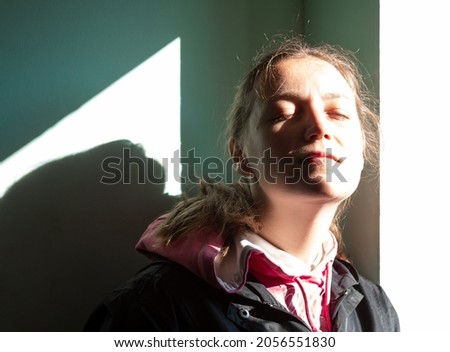 Happy young woman in black raincoat against wall basking in sun autumn day. Smiling girl with closed eyes standing near door sunrays. Lifestyle real people Seasonal depression mental health, Vitamin D