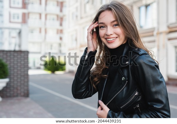 happy young woman in black\
turtleneck and leather jacket adjusting hair on urban street of\
europe