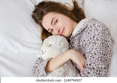 Happy Young Woman With Blonde Hair Sleeping In An Embrace With A Stuffed Animal Toy. Favorite Sheep In The Hands Of Teen Girl In Cute Warm Pajamas Close-up And Space On The Left. Top View. Copy Space.