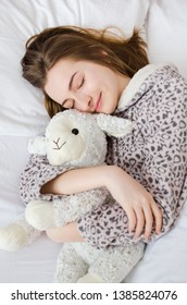 Happy Young Woman With Blonde Hair Sleeping In An Embrace With A Stuffed Animal Toy. Favorite Sheep In The Hands Of Teen Girl In Cute Warm Pajamas Close-up. Top View.