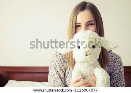Happy young woman with blonde hair sitting on the bed in her bedroom in an embrace with a stuffed animal toy. Favorite white sheep in the hands of teen girl in cute warm pajamas.