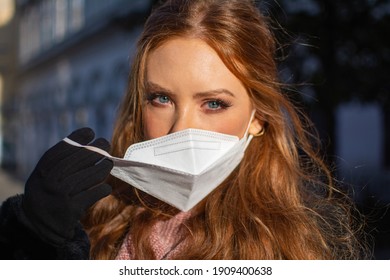 Happy Young Woman With Blue Eyes And Ginger Hair Removing A Protective Facial Mask (FFP-2) During The COVD-19 Pandemic Standing Outdoors On A Sunny Spring Day In A City Street Scene.