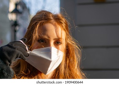 Happy Young Woman With Blue Eyes And Ginger Hair Removing A Protective Facial Mask (FFP-2) During The COVD-19 Pandemic Standing Outdoors On A Sunny Spring Day In A City Street Scene.