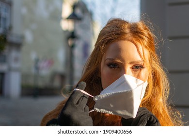 Happy Young Woman With Blue Eyes And Ginger Hair Removing A Protective Facial Mask (FFP-2) During The COVD-19 Pandemic Standing Outdoors On A Sunny Spring Day In A City Street Scene.