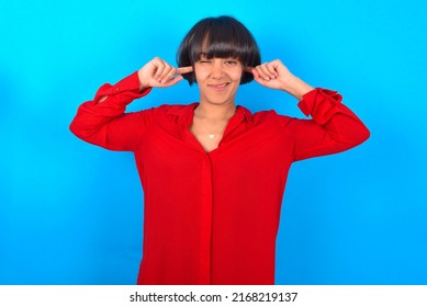 Happy Young Woman With Bob Haircut Wearing Red Shirt Over Blue Wall Ignores Loud Music And Plugs Ears With Fingers Asks To Turn Off Sound