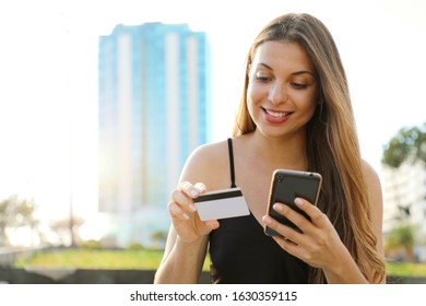 Happy Young Woman Booking Online Outdoor. Reservation Hotel With Smart Phone, Girl Reading Credit Card Number Outdoor On Summer.