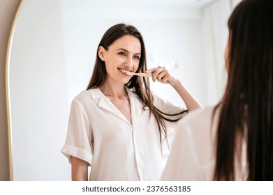 happy young woman brushing her healthy teeth with wooden toothbrush and whitening toothpaste while looking in mirror in bathroom interior. oral and dental care, morning routine concept - Powered by Shutterstock