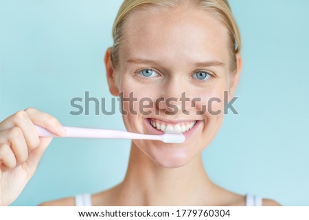 Happy young woman brushing her white teeth while looking at the camera satisfied with beautiful eyes. Isolated against a blue background.