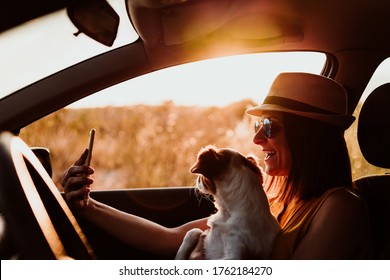 Happy Young Woman In A Car With Her Dog At Sunset. Taking A Picture With Mobile Phone. Travel Concept