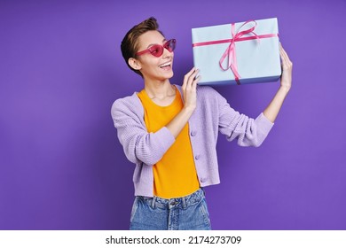 Happy Young Woman Carrying Gift Box On Shoulder While Standing Against Purple Background