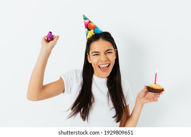 Happy Young Woman Celebrates Birthday Wearing Party Hat Holding Cake With Candle And Kazoo Standing On White Backdrop.