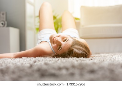 Happy Young Woman Chilling Out At Home Lying On Her Back On The Living Room Carpet Looking Back At The Camera In The Glow Of The Sun In A Low Angle View