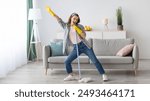 Happy young woman cleaning her home, singing at mop like at microphone and having fun, free space. Millennial housewife enjoying domestic chores, doing home cleanup creatively