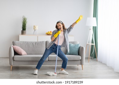 Happy young woman cleaning her home, singing at mop like at microphone and having fun, free space. Millennial housewife enjoying domestic chores, doing home cleanup creatively