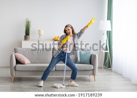 Happy young woman cleaning her home, singing at mop like at microphone and having fun, free space. Millennial housewife enjoying domestic chores, doing home cleanup creatively