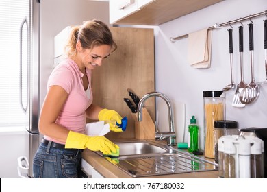 Happy Young Woman Cleaning Kitchen Sink With Sponge And Spray In Kitchen