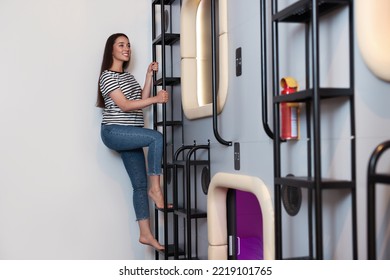 Happy Young Woman Climbing Up Ladder To Capsule In Pod Hostel