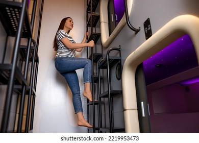 Happy Young Woman Climbing Up Ladder To Capsule In Pod Hostel, Low Angle View