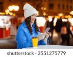 Happy young woman with coffee cup using cellphone outdoors at Christmas market, festive holidays background, free space