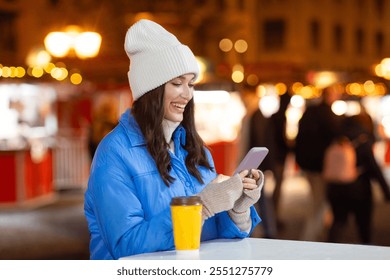 Happy young woman with coffee cup using cellphone outdoors at Christmas market, festive holidays background, free space - Powered by Shutterstock