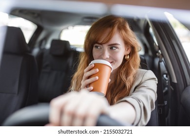 Happy young woman with coffee to go driving her car. Woman sipping a coffee while driving a car. Young woman drinking coffee while driving her car. Attractive red hair drives a car - Powered by Shutterstock