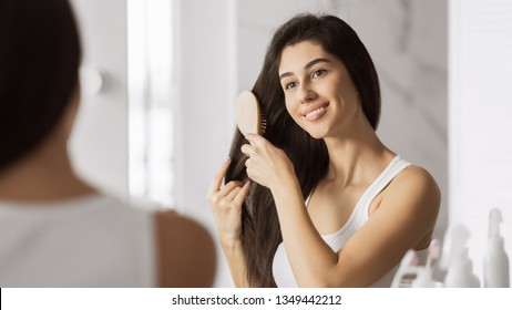 Happy Young Woman Combing Her Hair And Smiling Looking At The Mirror. Everyday Haircare Concept