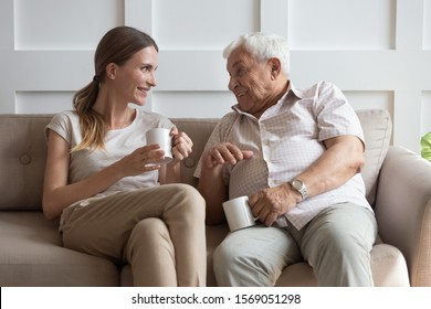 Happy young woman communicating with smiling old father, sitting together on comfortable couch in living room. Excited retired 70s man talking over cup of coffee tea with joyful grown up daughter.