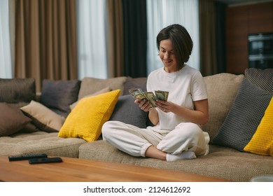 Happy young woman counting cash, enjoying money win, big profit, salary in payday sitting on sofa at home