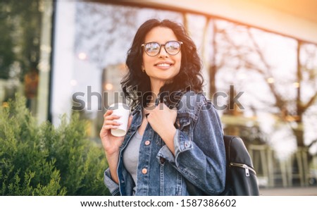 Happy young woman with cup of drink to go smiling and looking away while walking on city street on sunny day