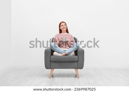Happy young woman with cup of drink sitting in armchair near white wall indoors