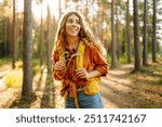 A happy young woman with curly hair walks along a forest path, wearing a yellow hiking backpack surrounded by tall trees and warm sunlight