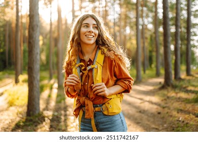 A happy young woman with curly hair walks along a forest path, wearing a yellow hiking backpack surrounded by tall trees and warm sunlight - Powered by Shutterstock