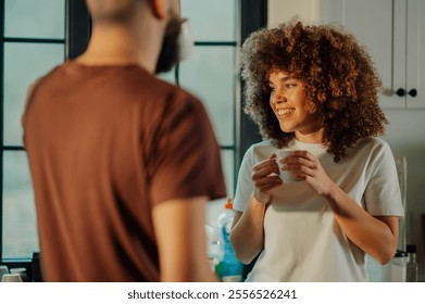 Happy young woman with curly hair holding a coffee cup and smiling at her partner in a sunlit kitchen, enjoying a quiet morning moment together - Powered by Shutterstock