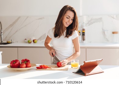 Happy young woman cutting vegetables for the salad from a recipe and looking at the tablet computer in a kitchen - Powered by Shutterstock