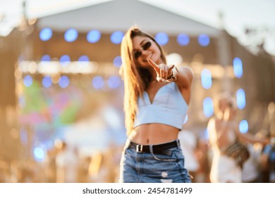 Happy young woman dances at beach music fest, summer vibe. Casual outfit, sunlight, crowd. Lady enjoys live concert, points, smiles. Youth lifestyle, entertainment begins at outdoor event. - Powered by Shutterstock