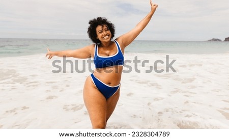 Happy young woman dancing and celebrating at the beach, wearing a bikini and raising her arms in excitement. Carefree, plus size woman having fun on a solo summer vacation.