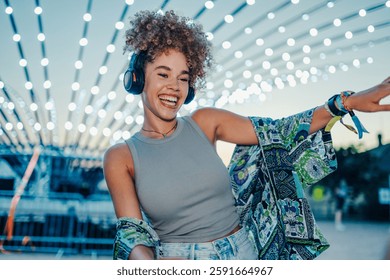Mujer joven feliz que baila alegremente mientras que lleva los auriculares inalámbricos, empapando la atmósfera vibrante de un festival de verano iluminado por luces decorativas coloridas