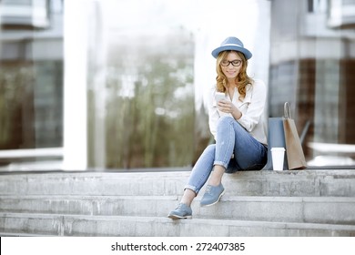 Fröhliche junge Frau mit einem Einwegkaffee und Einkaufstüten, die auf der Treppe sitzen und ihr Smartphone für die Kommunikation über Wi-Fi Internet in einem Sommerpark benutzen.