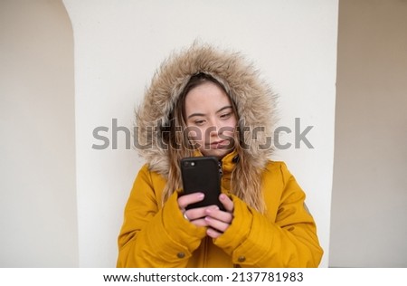Happy young woman with Down syndrome weraing parka, leaning the wall and using smartphone.