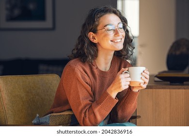 Happy young woman drinking a cup of tea in an autumn morning. Dreaming girl sitting in living room with cup of hot coffee enjoying under blanket with closed eyes. Pretty woman wearing sweater at home. - Shutterstock ID 2042836916