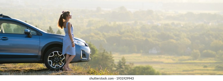 Fröhliche junge Frau Fahrer in blauem Kleid genießen warmen Sommerabend neben ihrem Auto stehen. Reise- und Urlaubskonzept.