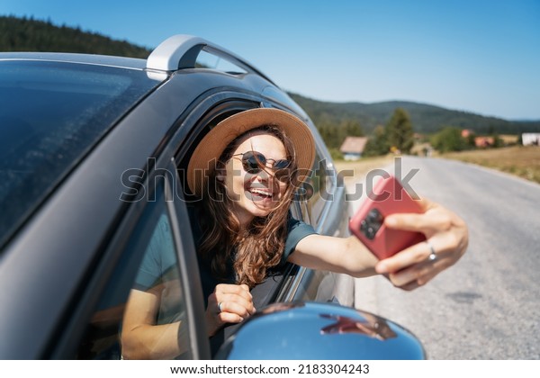 Happy\
young woman driver traveler in hat and sunglasses sitting behind\
the wheel of car taking selfie using\
smartphone